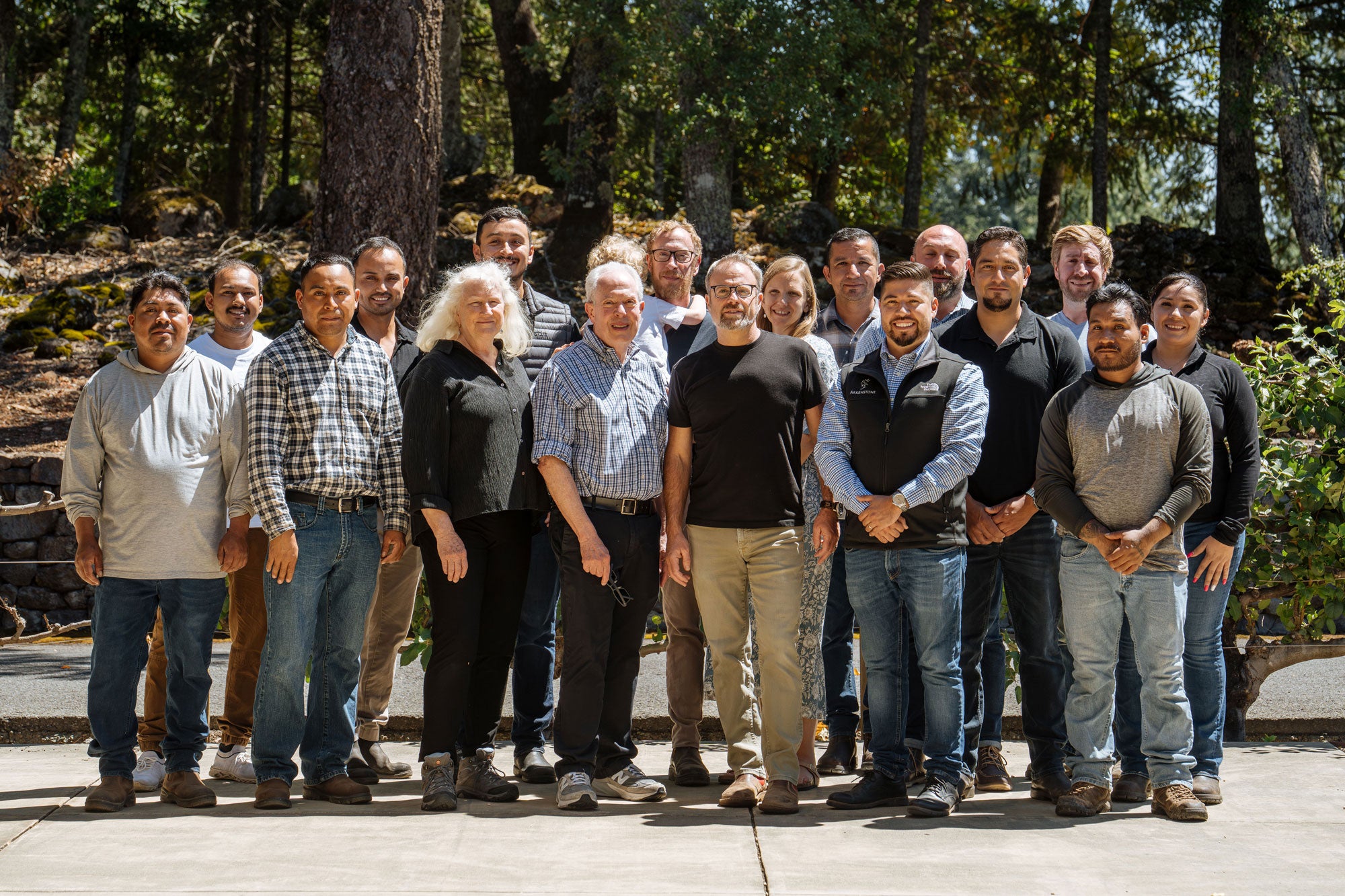 A group of nineteen people stands outdoors on a sunny day, smiling at the camera. They are positioned in front of trees and greenery, standing on a paved area. Most are casually dressed.