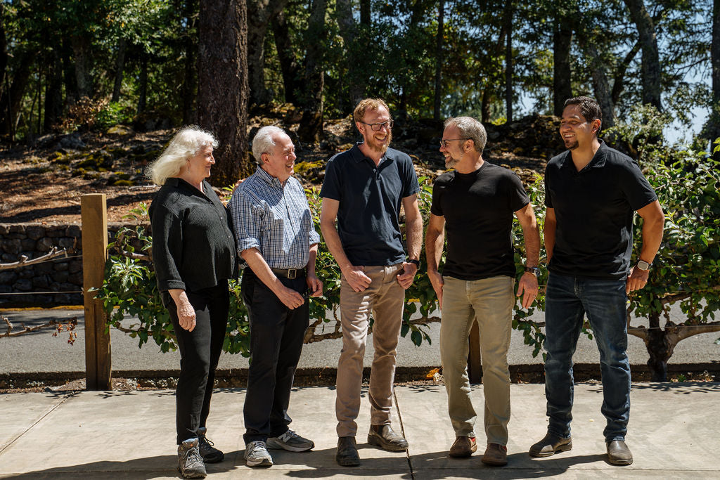 Five adults stand outdoors on a sunny day, smiling and talking together. They are casually dressed, with trees, greenery, and a wooden fence in the background.