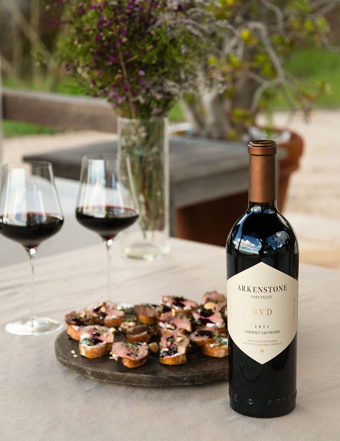 A bottle of Arkenstone NVD Cabernet Sauvignon stands on a table with two glasses of red wine and a plate of small appetizers, set outdoors with a blurred natural background.