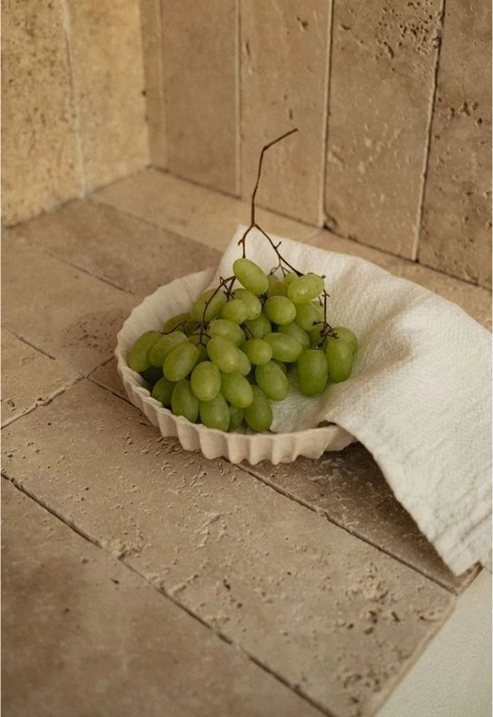 A bunch of green grapes rests on a white cloth inside a scalloped ceramic dish, placed on a beige stone tile surface against a matching tiled wall.