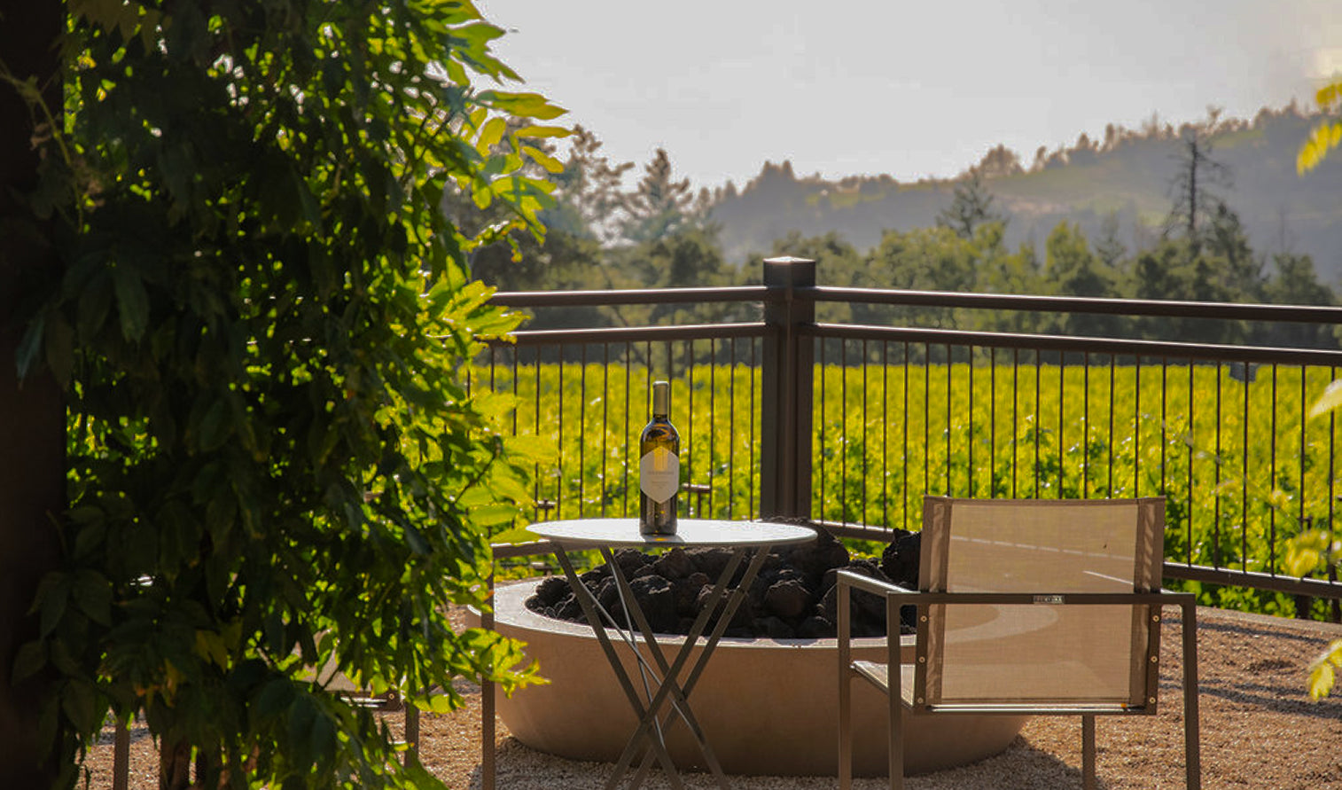 A patio with a metal table, a bottle of wine, and two chairs overlooks a lush green vineyard and rolling hills under a bright sky, with sunlight filtering through nearby trees.