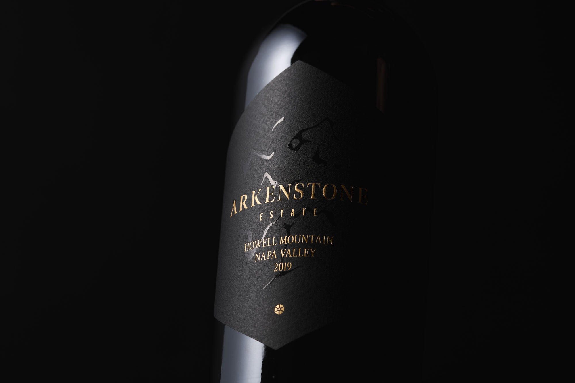 A close-up of a black wine bottle with a black and gold label reading Arkenstone Estate, Howell Mountain, Napa Valley, 2019 against a dark background.