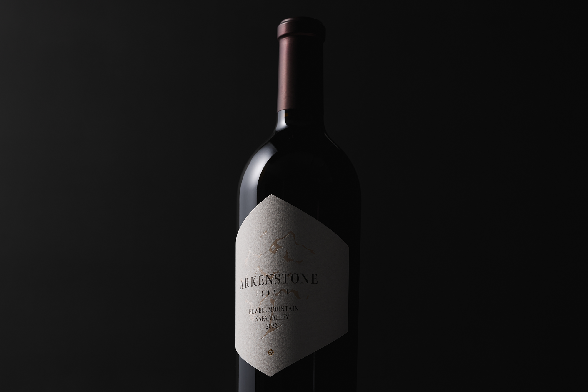 A dark wine bottle of 2023 Estate Red, with a maroon foil top and cream label, is set against a black background and partially lit from the left.