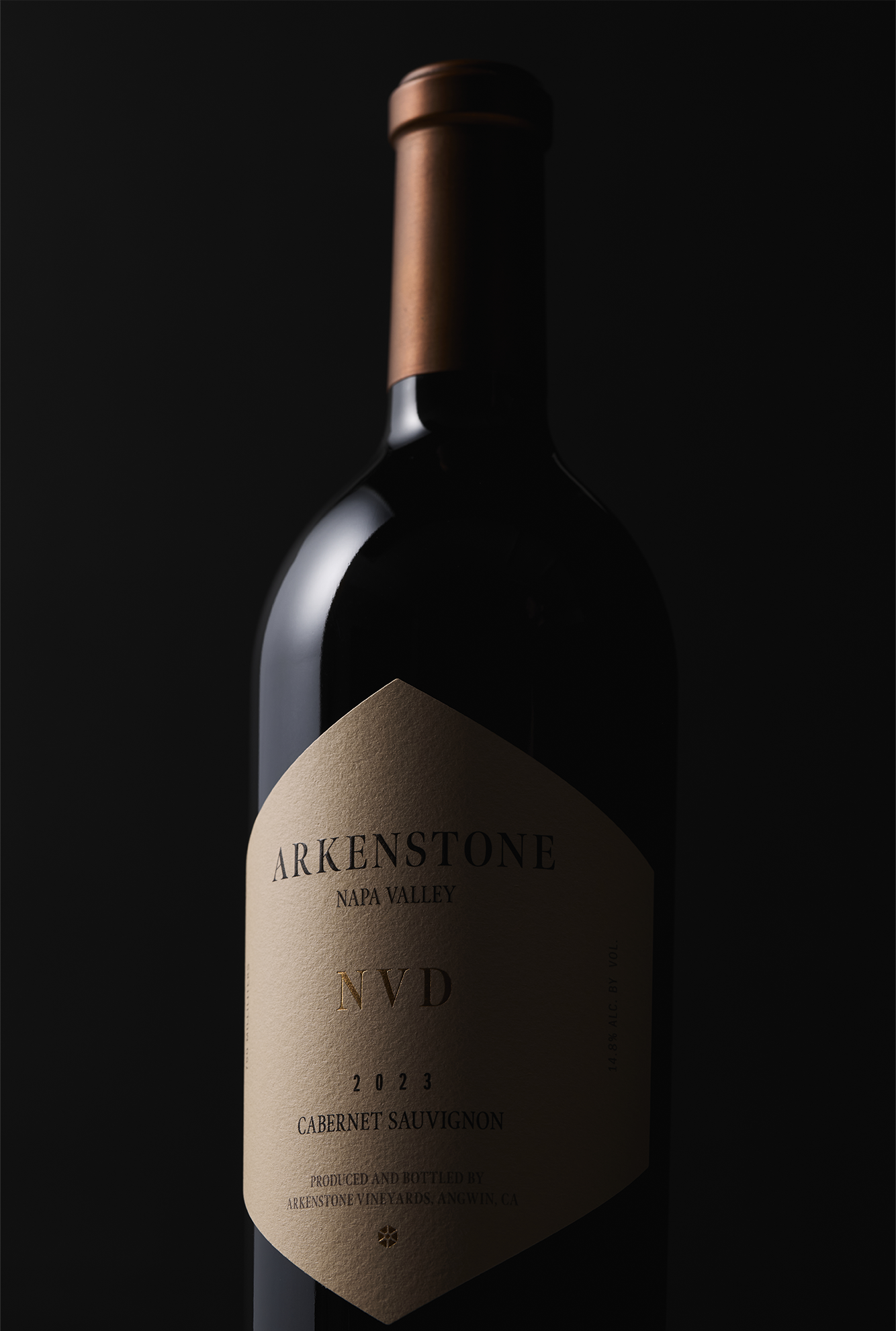 A bottle of 2023 NVD with a tan label and copper foil cap stands against a dark, dramatic background. This Howell Mountain estate Cabernet blend wine exudes elegance and depth.